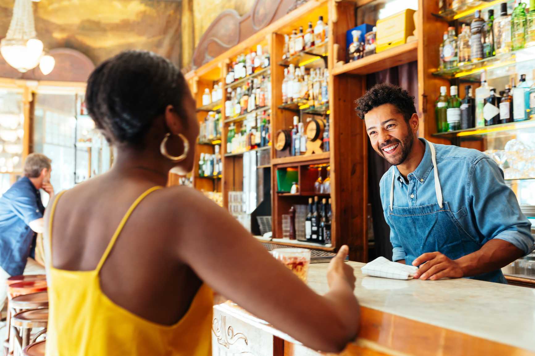 Cheerful ethnic barmen in denim uniform smiling and speaking with female customer while working in cozy bar
